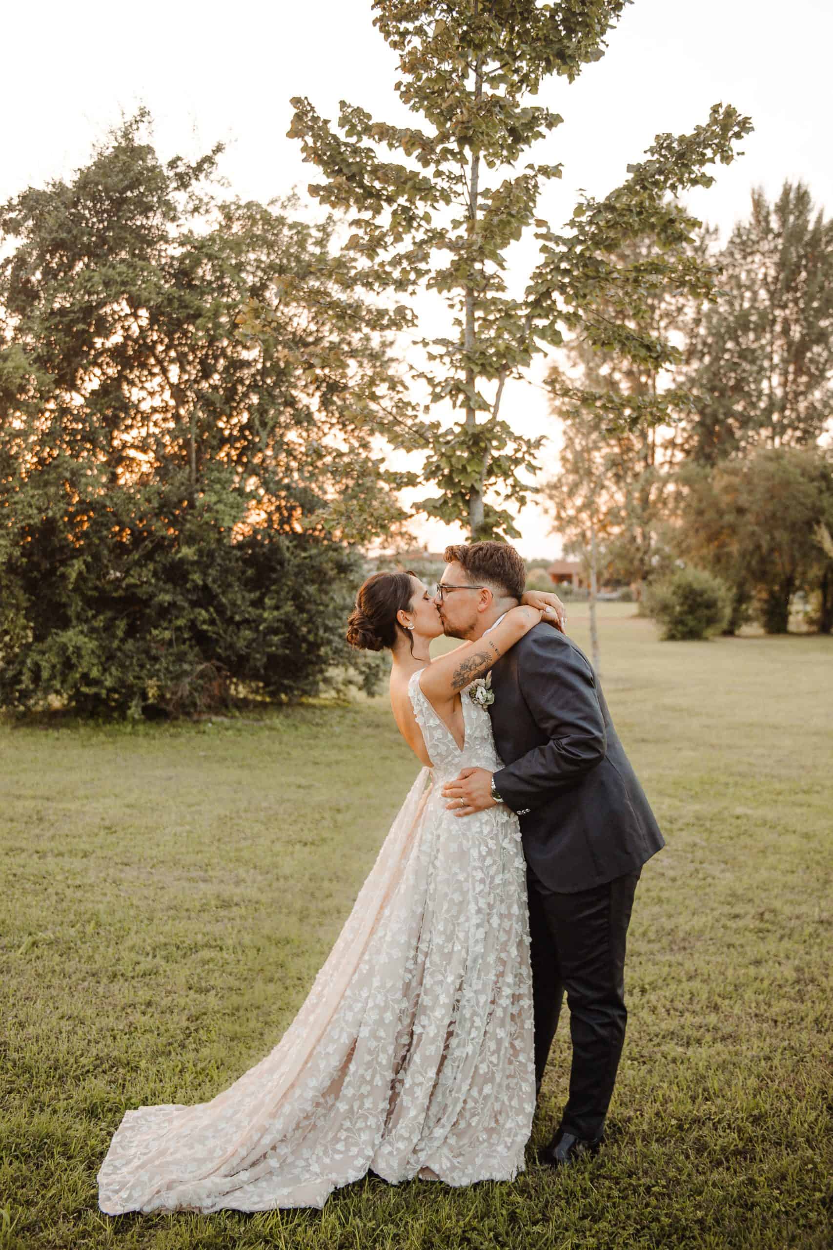 vediamo una sposa e uno sposo che si baciano e si tengono di lato, in un bellissimo campo di erba verde e alberi. lei indossa un bellissimo abito da sposa fatto su misura e lui indossa un abito