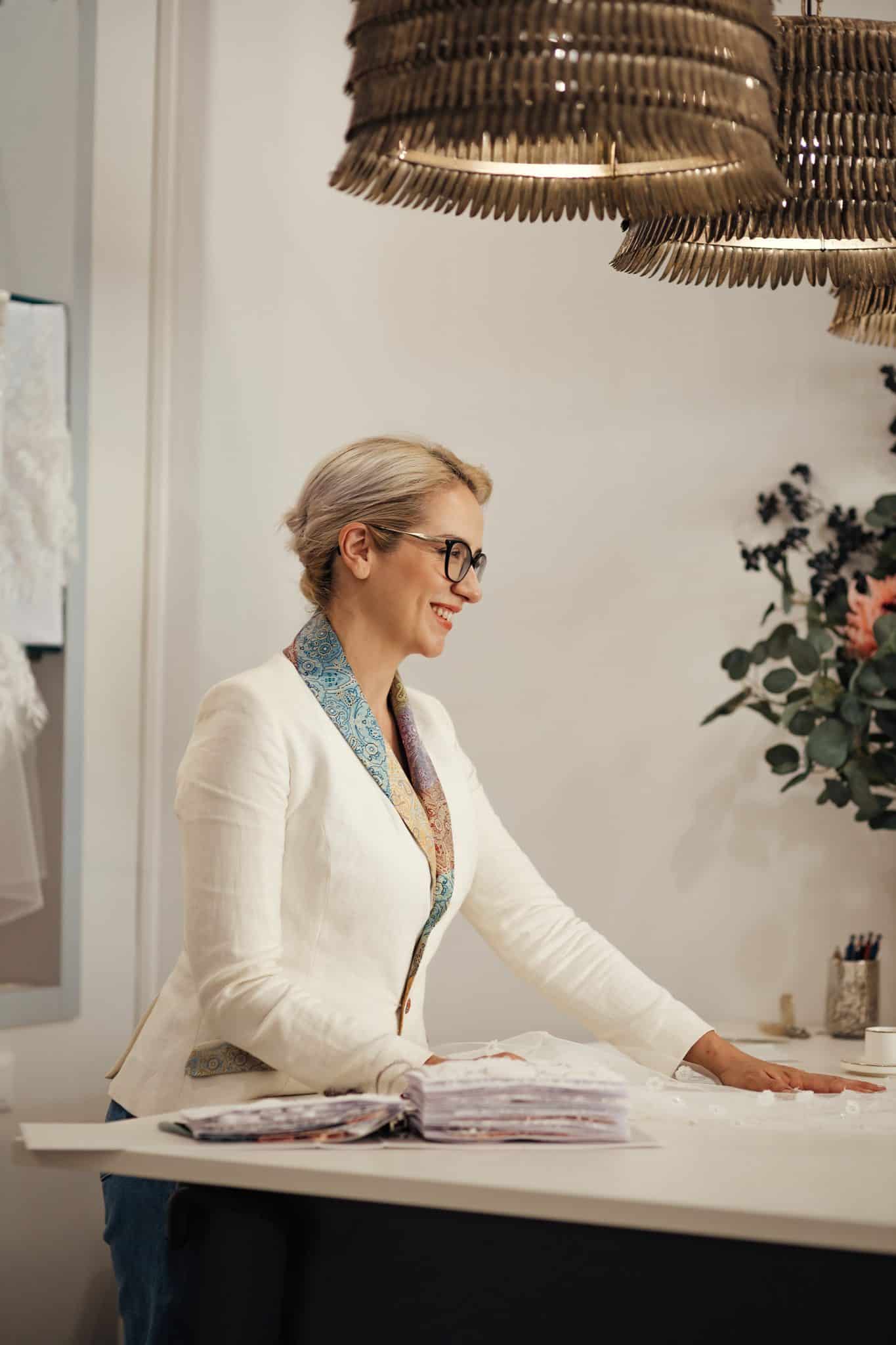 A blonde woman, designer Angelina Haole, is turned towards the right side of the frame, smiling, with her hands on a work surface. She wears a white linen sport coat with a colorful lapel and there are gold lamps over her head and flowers on the table. her hair is pulled back into a chignon. 