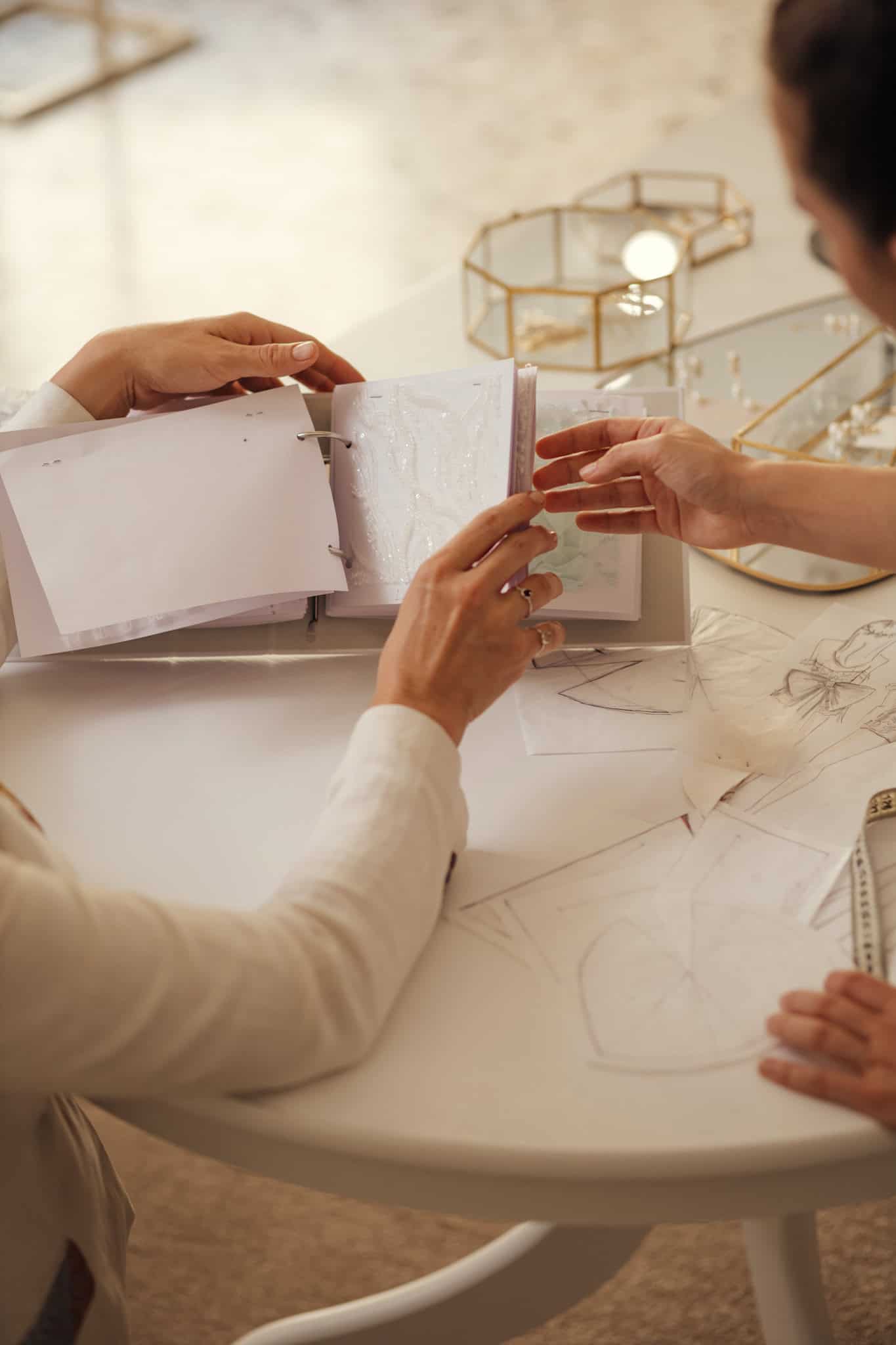 We see a white table with fashion illustrations, pattern pieces and fabric samples. Two sets of hands are touching a fabric sample in what seems to be a selection process