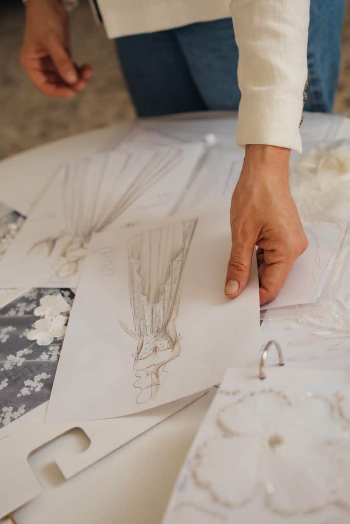 La mano della stilista Angelina Haole è mostrata mentre tiene un'illustrazione di moda sopra un tavolo con pezzi di cartamodelli, campioni di tessuto e altri disegni. il contrario di abiti da sposa fast fashion