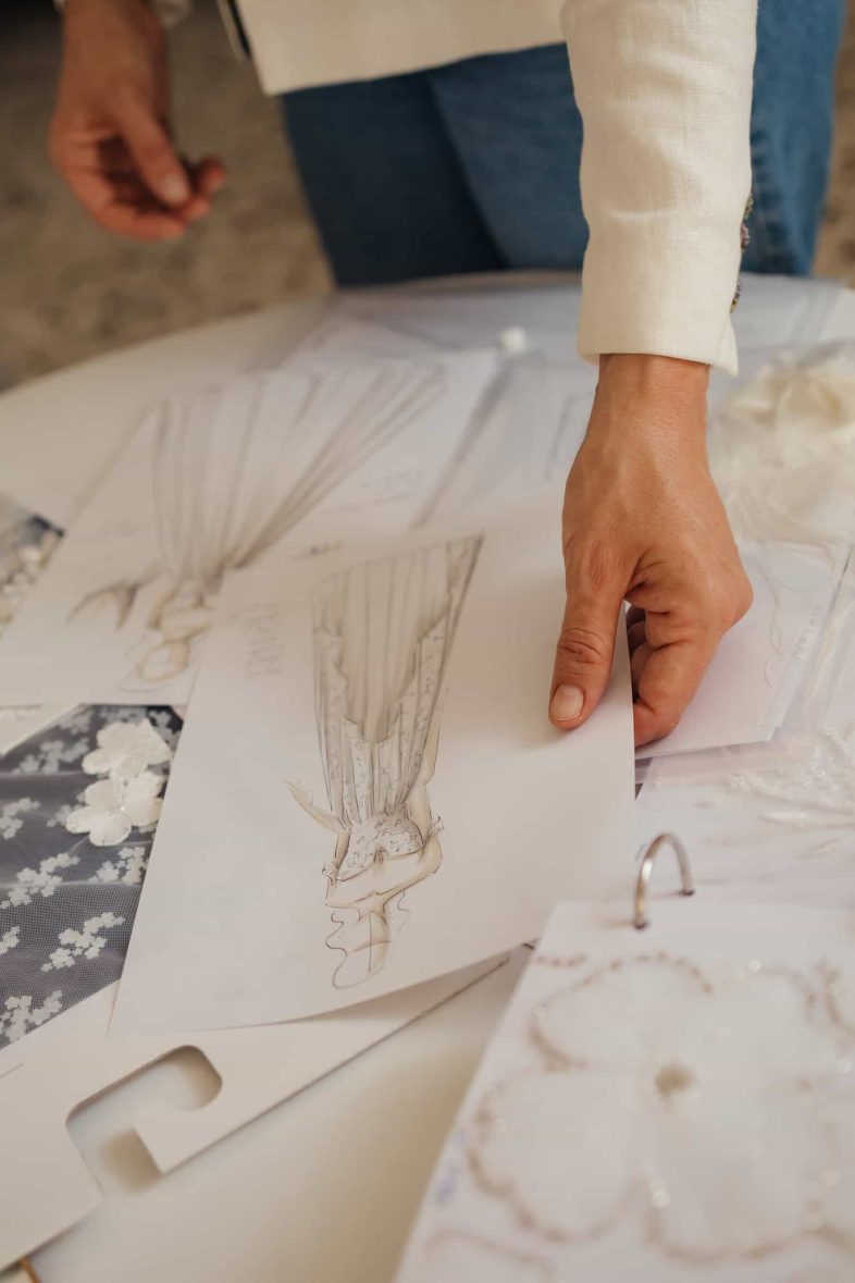 La mano della stilista Angelina Haole è mostrata mentre tiene un'illustrazione di moda sopra un tavolo con pezzi di cartamodelli, campioni di tessuto e altri disegni.contatta atelier sposa Soave